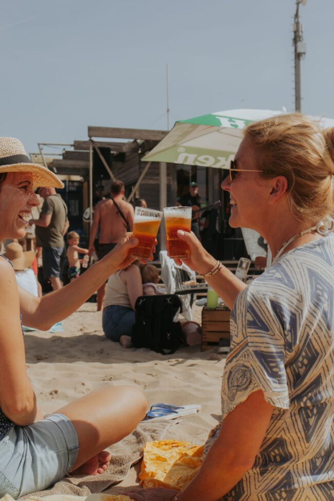 Vriendinnen genieten van een zonnige dag op het strand met een drankje in de hand, omringd door een levendige sfeer.