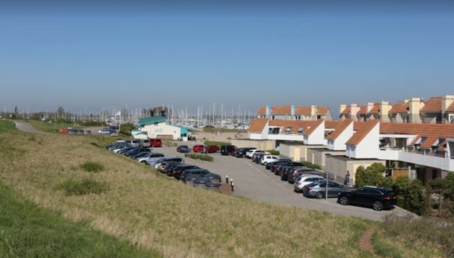 Parkeerterrein vol met auto's naast een grasveld met op de achtergrond huizen en masten van zeilboten in Brouwershaven, Zeeland (2)