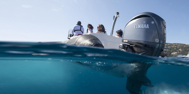 Achterkant van een motorboot met Yamaha buitenboordmotor op helderblauw water, met passagiers aan boord aan de kustlijn