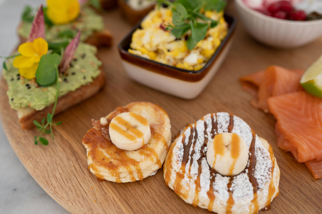 Houten serveerplank met diverse ontbijtgerechten, waaronder toast met avocado, roerei, en pannenkoeken bij Liefs Lies in Breskens