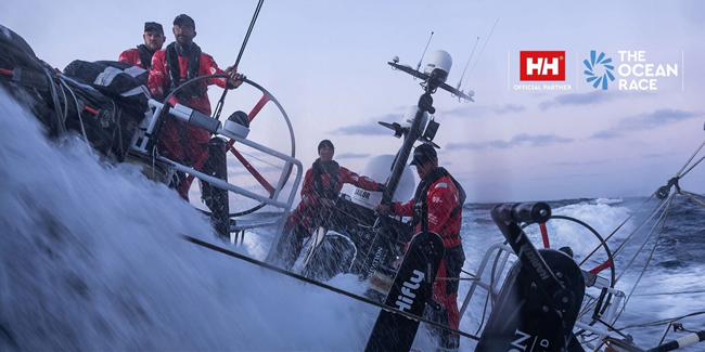 Zeilers in waterdichte kleding besturen een zeilboot tijdens een stormachtige zeilrace, met logo's van HH en The Ocean Race zichtbaar.