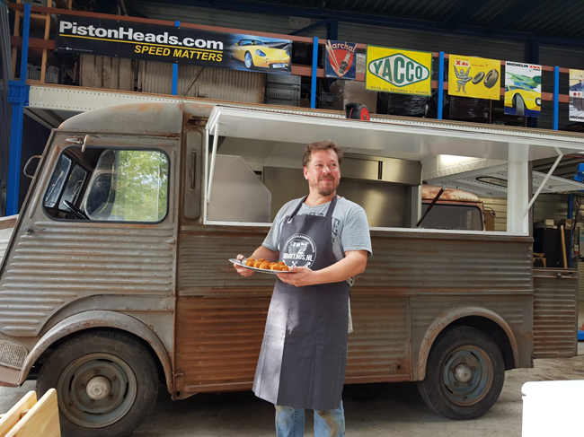 Chef presenteert een bord met heerlijke snacks voor een vintage Citroën HY foodtruck in een garage.