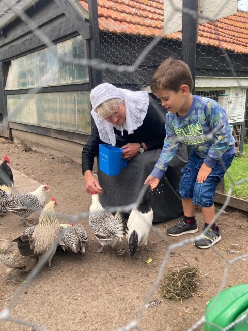 kind en oudere vrouw in traditionele klederdracht voeren samen kippen op een boerderij