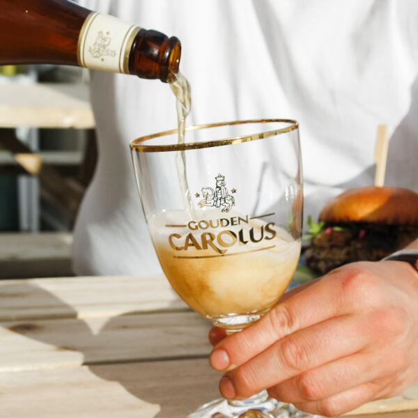 Bier wordt ingeschonken in een Gouden Carolus-glas dat door een hand wordt vastgehouden, burger op de achtergrond bij Strandp