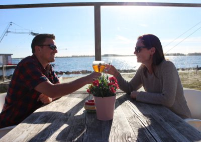 Man en vrouw aan houten tafel op terras bij water proosten met glazen bier, plant op tafel bij Restaurant & Terras Beau4