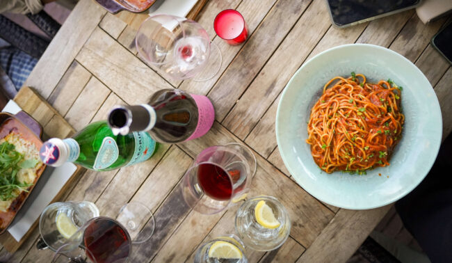 Bovenaanzicht van houten tafel met bord spaghetti in tomatensaus, glazen rode wijn, flessen en water met citroen, bij Pizzeri