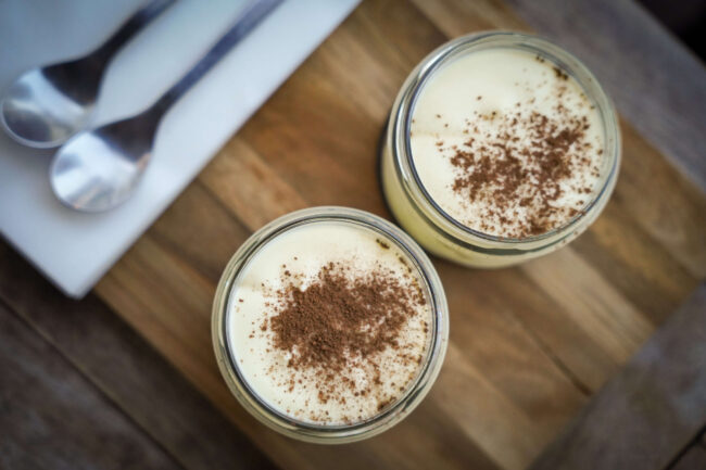 Twee glazen potjes met romig dessert bestrooid met cacao op een houten plank, naast twee lepels bij Pizzeria Trattoria Napoli