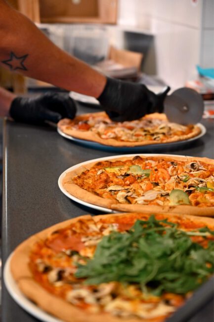 Hand met zwarte handschoen snijdt pizza met pizzasnijder; op de voorgrond borden met pizza's en groenten, bij Napoli