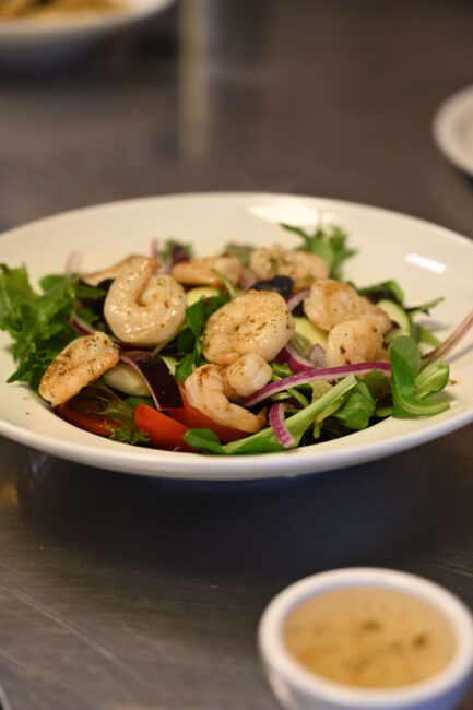 Wit bord met salade van gemengde sla, tomaat, rode ui en gebakken garnalen, met klein sausbakje op de voorgrond bij Pizzeria