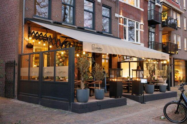Terras met overkapping, warme verlichting, planten en buitenhaarden voor een bakstenen gevel, bij Samen Oisterwijk