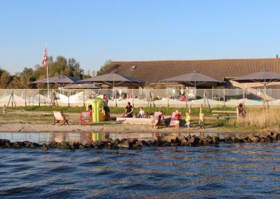 Waterkant met klein zandstrand en spelende kinderen, parasols en terras bij een laag gebouw, bij Restaurant & Terras Beau4