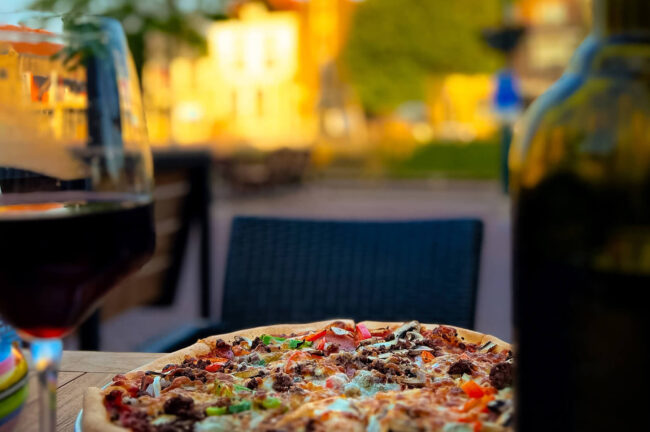 Close-up van pizza met kaas, vlees en paprika, met glas donker drankje en vervaagde achtergrond bij Pizzeria Trattoria Napoli