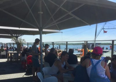 Buiten terras met grote parasols, mensen zittend en enkele staand aan tafels, uitzicht op het water en boten, bij Restaurant