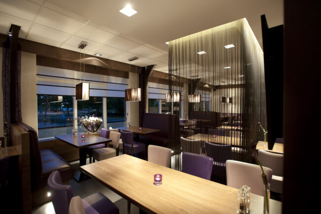 Interieur van modern restaurant met houten tafels, paarse en beige stoelen, hanglampen en een gordijn van fijne draden als sc