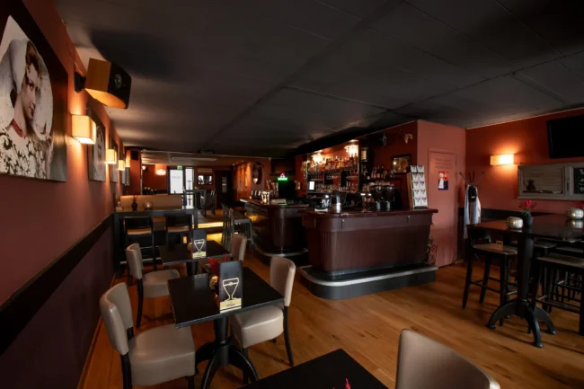 Interieur van een donker eetcafé met tafels en stoelen, houten bar met flessenrekken en warme wandlampen bij Hart van Tholen