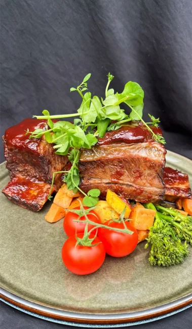 Gegrilde ribstukken met glazuur, jonge blaadjes, tros cherrytomaatjes en blokjes oranje groente op bord bij Hart van Tholen