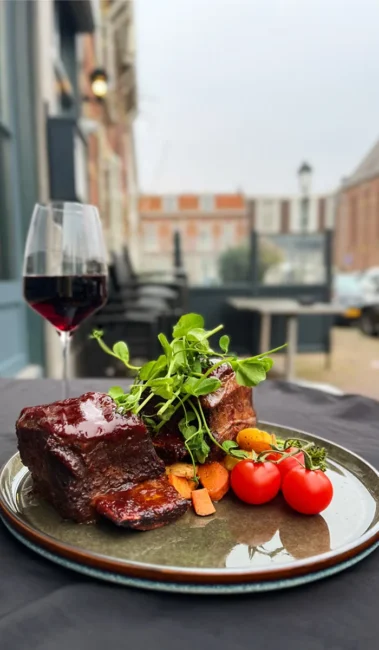 Bord met glanzend vlees, geroosterde wortelblokjes, cherrytomaten en verse kiemen op buitenterras, glas rode wijn, bij Eetcaf