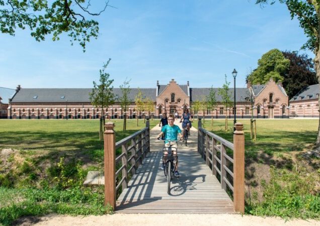 Fietsers genieten van een zonnige dag terwijl ze over een houten brug naar een historisch gebouw rijden.