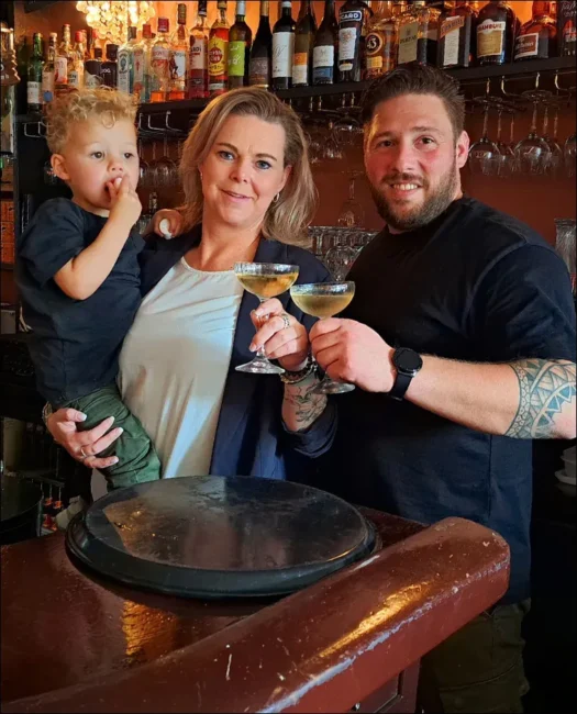 Vrouw die een peuter vasthoudt en een man die samen twee gevulde coupeglazen proosten voor een bar met flessen, bij Eetcafé H