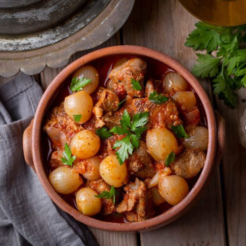 Terracotta kom met gestoofd vlees en pareluitjes in tomatensaus, gegarneerd met peterselie op houten tafel